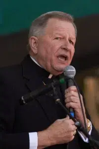 New Orleans archbishop speaking into a handheld microphone during a public event, wearing clerical clothing and a black suit jacket.