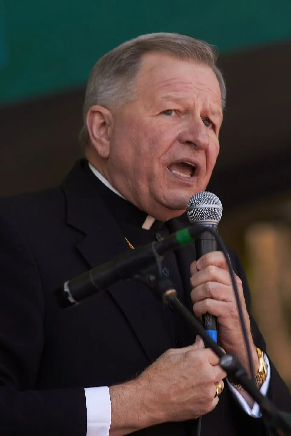 New Orleans archbishop speaking into a handheld microphone during a public event, wearing clerical clothing and a black suit jacket.