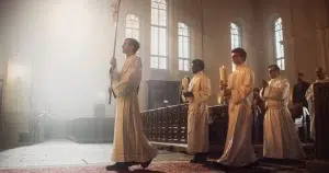 Clergy members in white robes processing down the aisle of a church, carrying candles and a cross, with soft light streaming through large stained glass windows.