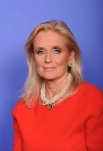 Official portrait of Congresswoman Debbie Dingell wearing a red dress and pearl necklace, posing against a blue studio background.