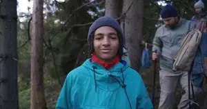 Teen wearing a jacket and beanie standing in a forest while staff members walk behind carrying backpacks, representing a troubled teen wilderness program setting