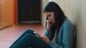 Teen girl sitting on the floor in a hallway, looking down at a smartphone with a worried and emotional expression.