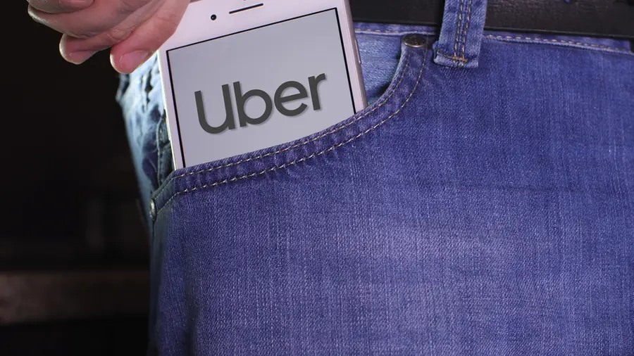 Smartphone displaying Uber logo being placed into a jeans pocket, symbolizing rideshare use and safety concerns.
