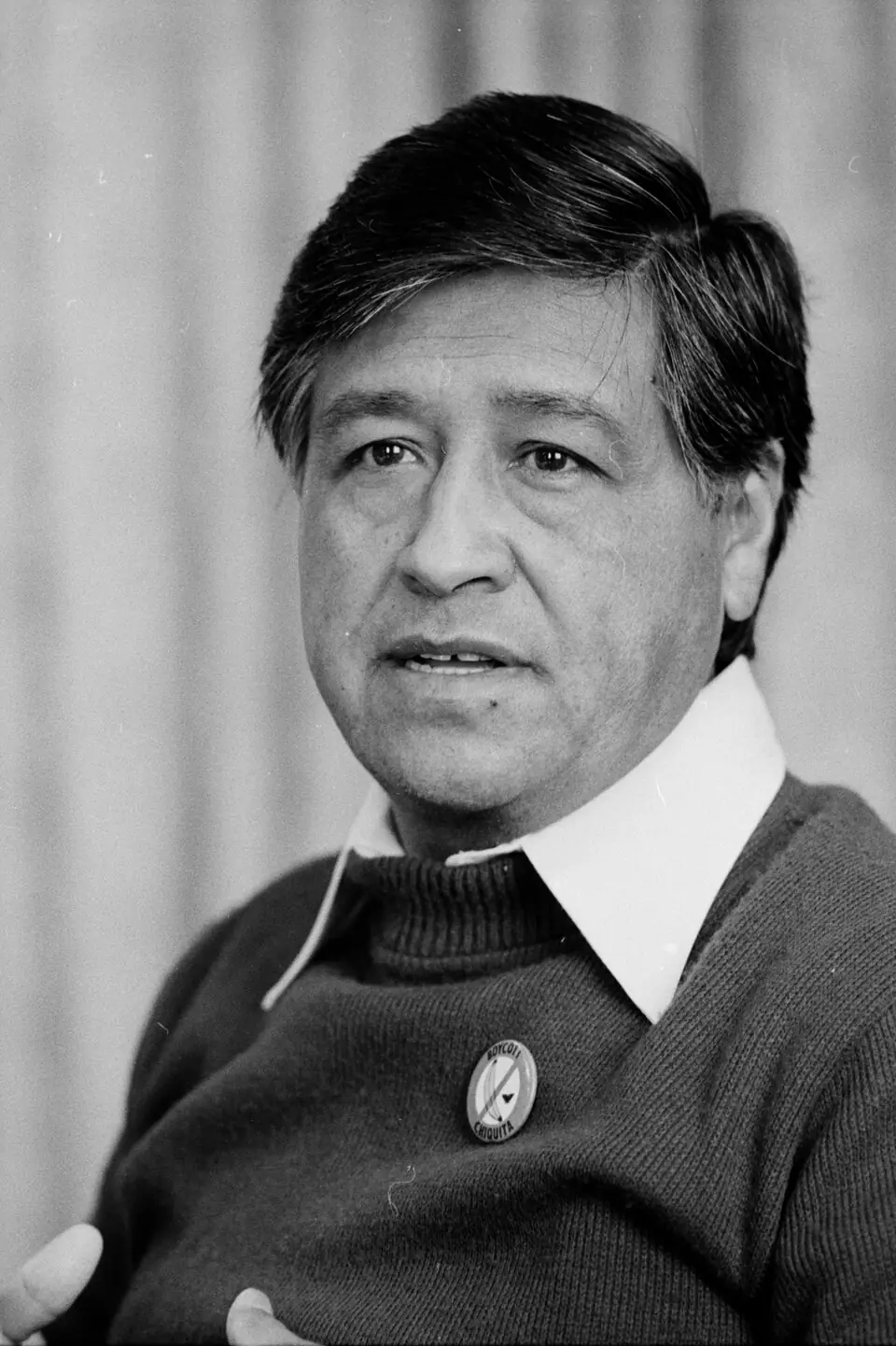 Black-and-white portrait of Cesar Chavez wearing a sweater and collared shirt, speaking with hands slightly raised, against a plain curtain background.