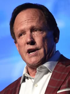 Jeff Webb speaking at an event, wearing a red plaid jacket and white shirt against a blue background.