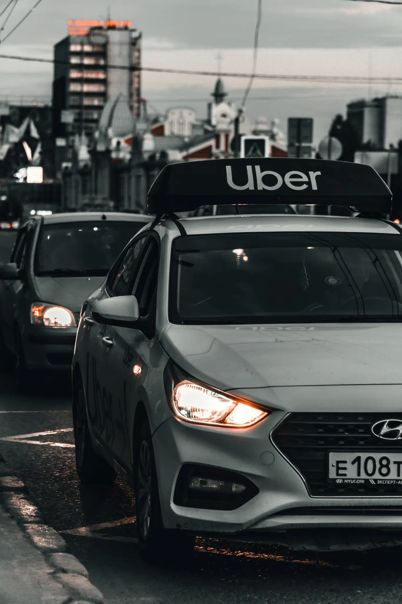 Uber-branded vehicle in traffic on a city street at dusk with headlights on and urban buildings in the background