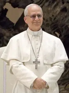 pope-leo-xiv Pope Leo XIV wearing white papal vestments and a silver cross necklace during a public appearance.